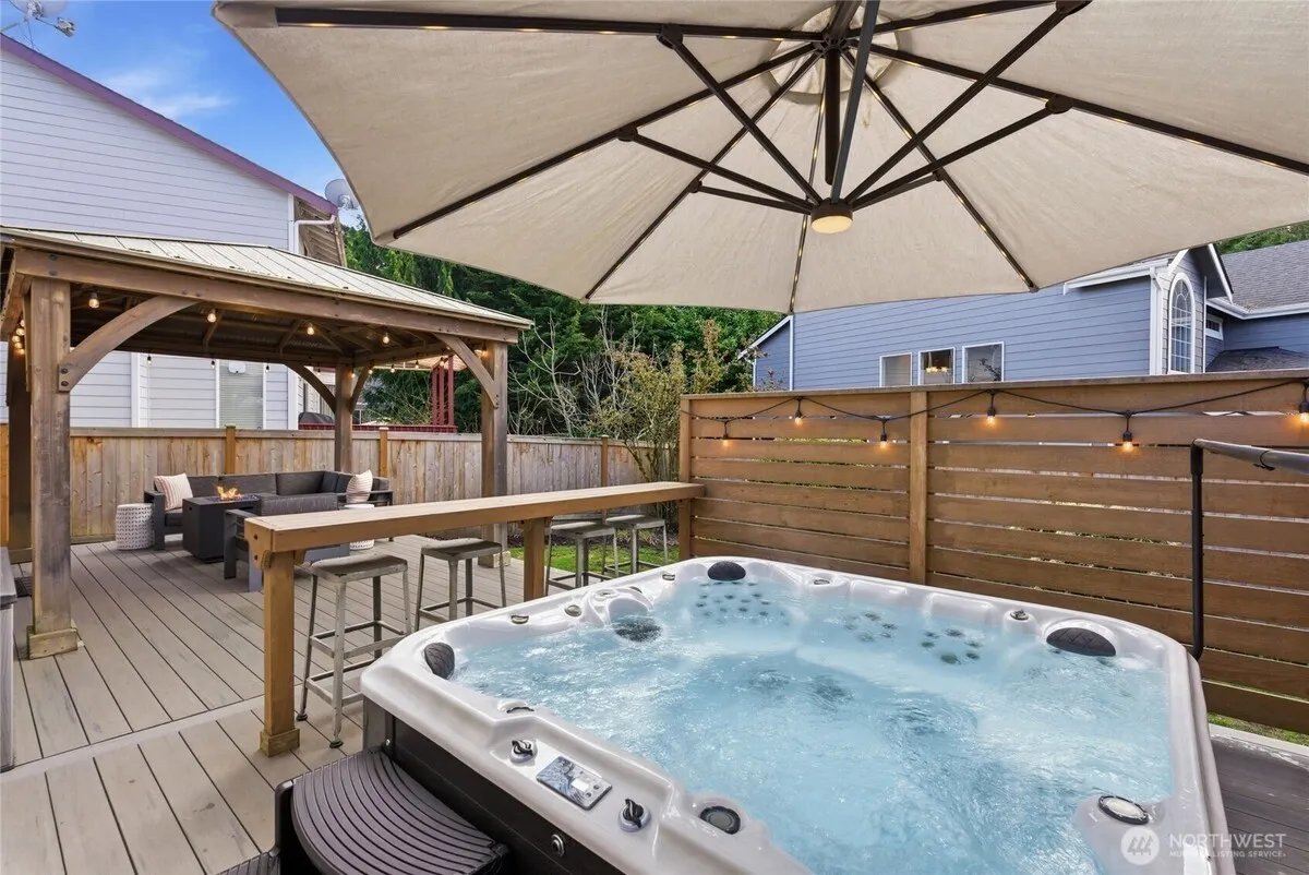 A premium hot tub on a deck under a large parasol, with a small slatted wood privacy screen behind it — and two neighboring houses clearly visible above